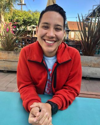 photo of author Lourdes Rivas sitting outside at a blue table wearing a red coat and smiling