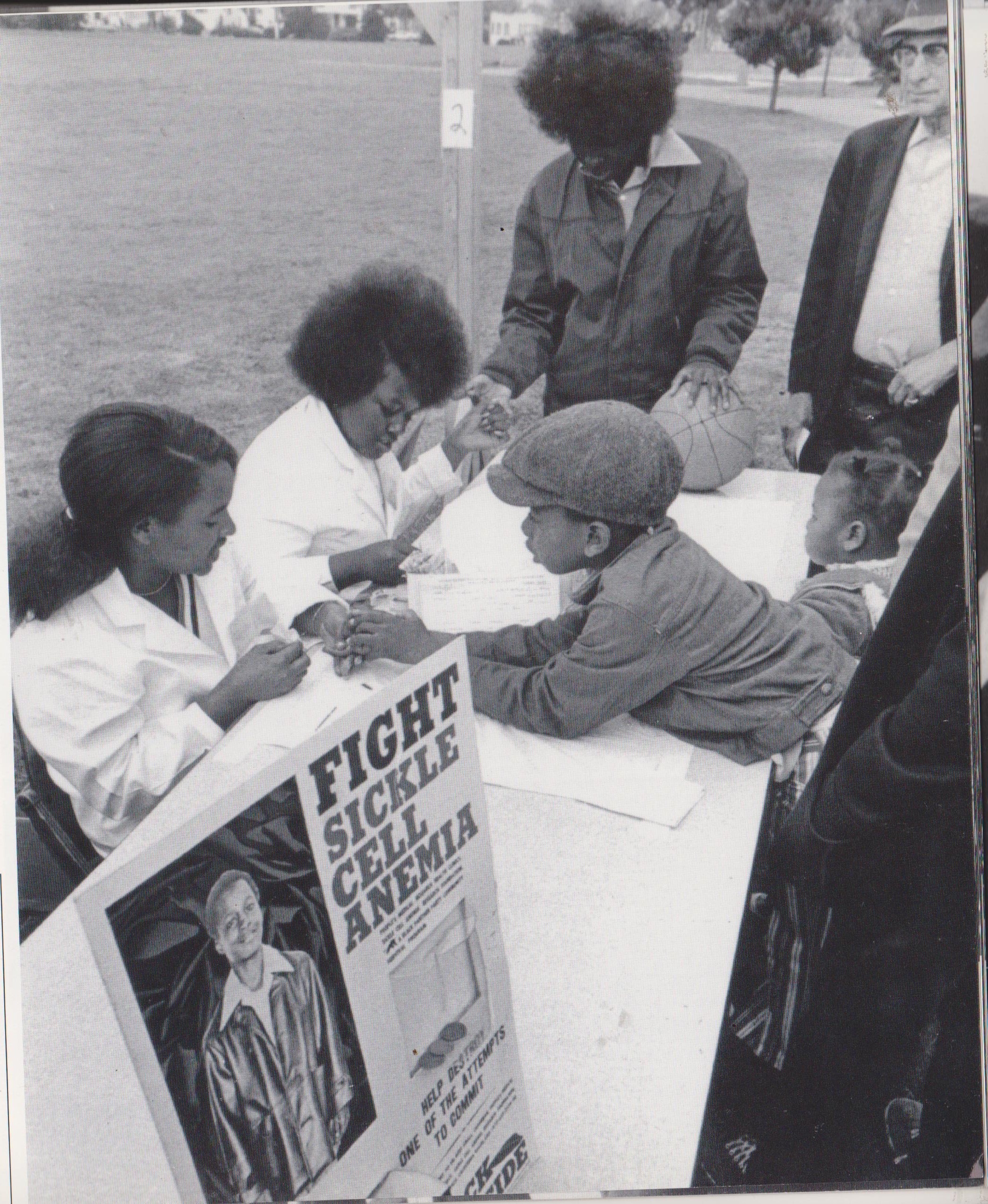 Black and white photograph of children being seen by nurses at a table in a public park during a community event
