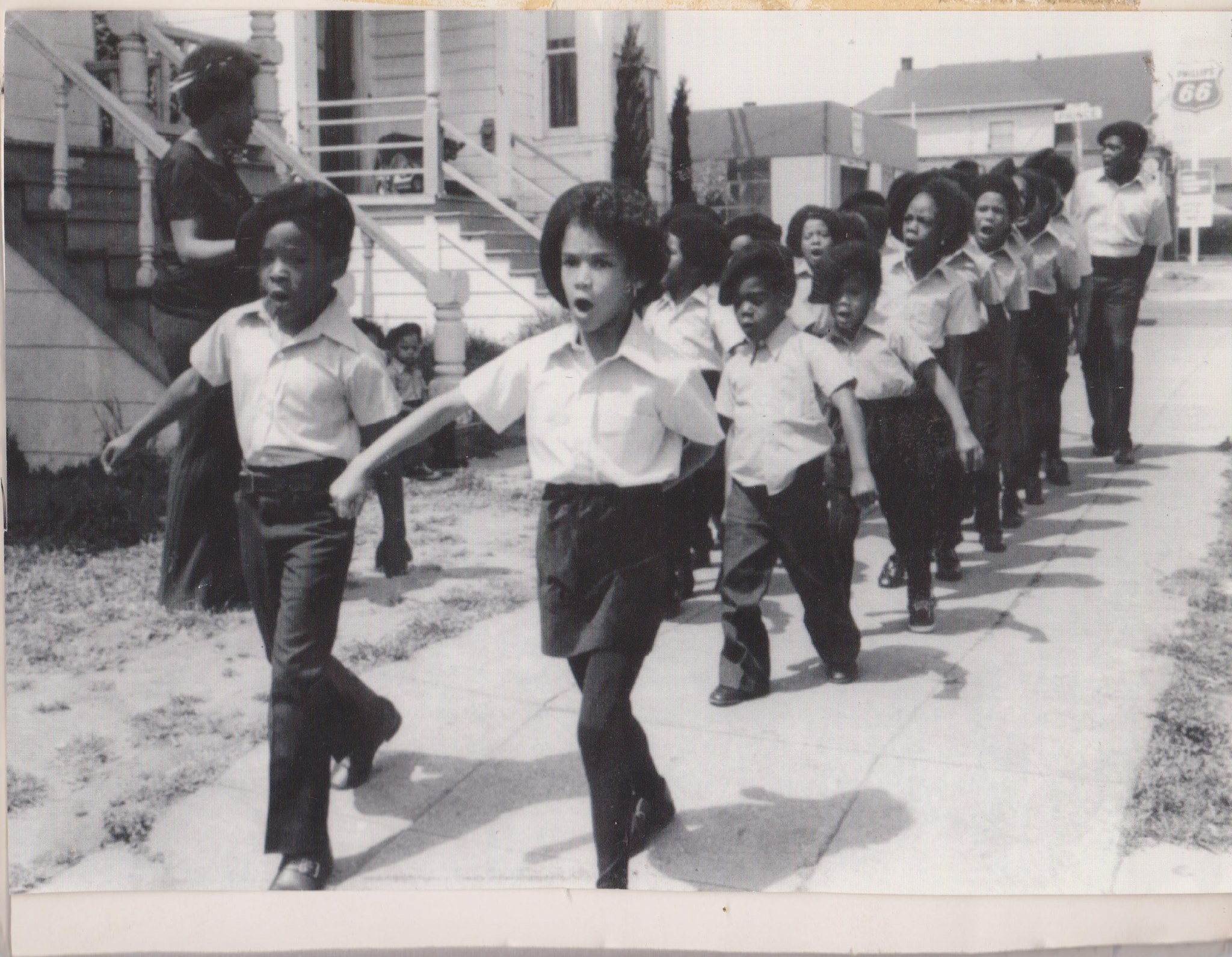 Black and white photograph of a group of school children marching down a residential street in unison. The children are wearing a uniform of black pants, white shirts, and black berets.