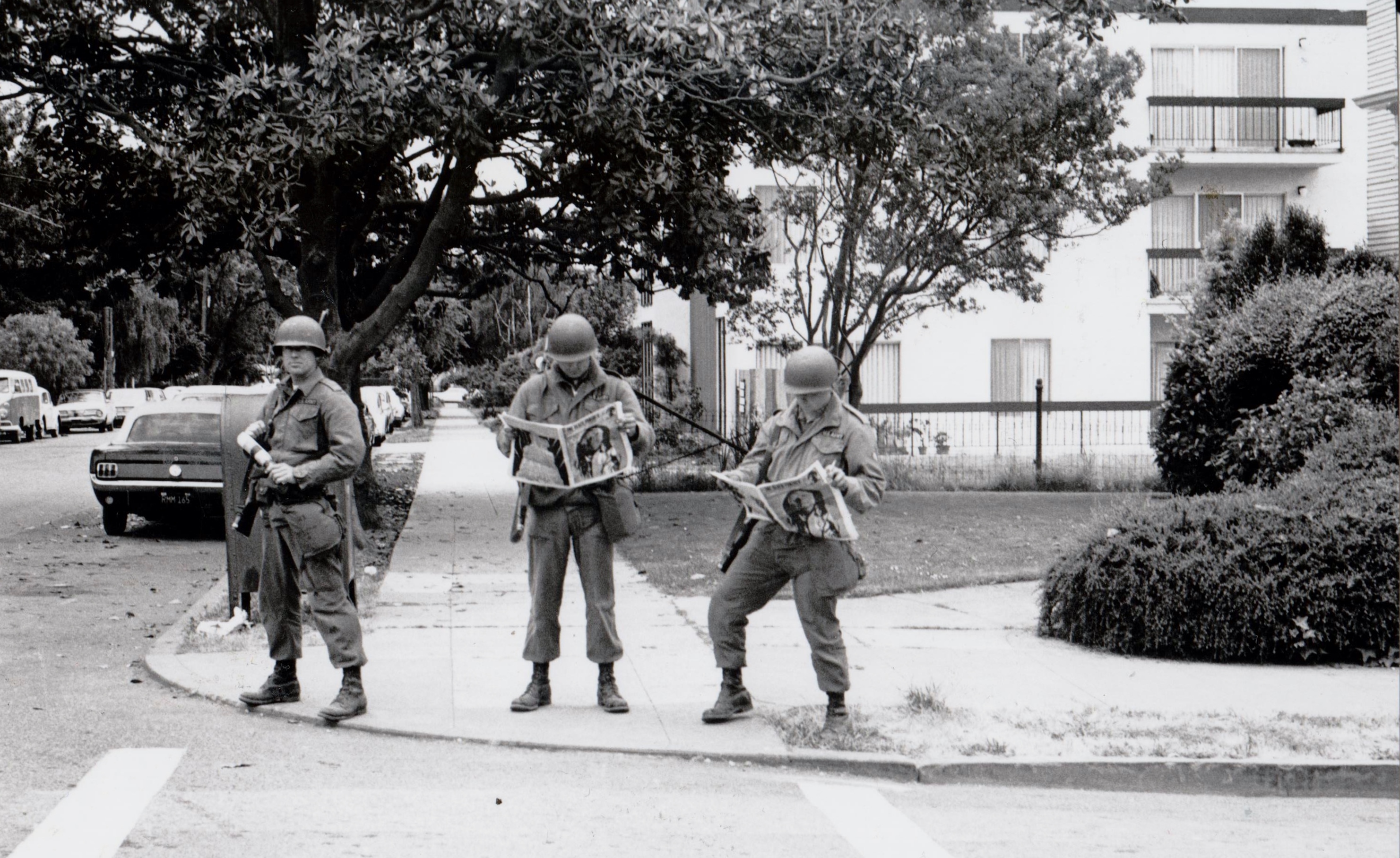 Black and white photograph of three national guardsmen in full gear reading a newspaper on a residential street corner