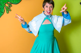Risa in front of an orange and green wall, wearing a blue dress with a cape