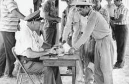 Man at a table outside with another man behind it with men in background