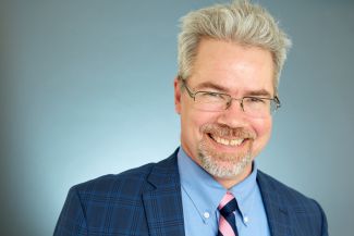 Headshot of Henry, smiling, wearing glasses and a suit with tie