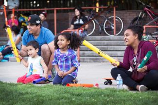 photo of man and child playing with yellow plastic drumstick, girl sitting nearby and woman holding another yellow plastic drumstick