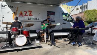 White van with the words "Bay Area Jazz Mobile" with three musicians in front playing drums, guitar and keyboards