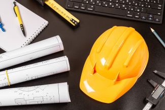 photo of yellow hard hat on a black desk with blueprints and computer keyboard
