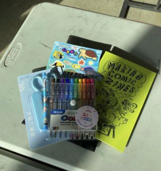 tabletop with a stack of zine supplies like pens/markers, stickers, scissors, and a finished zine