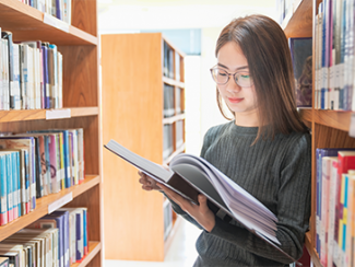 Woman standing next to a book shelf and looking at an open book