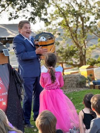photo of Germar the Magician outdoors holding a wooden bucket with a bunny in it. A child from the audience wearing pink is standing up facing the bunny. 