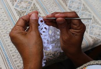 Two black hands holding a small crochet hook, making a doilie