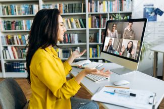 Woman in yellow talking on Zoom call with 4 other people on the computer screen.