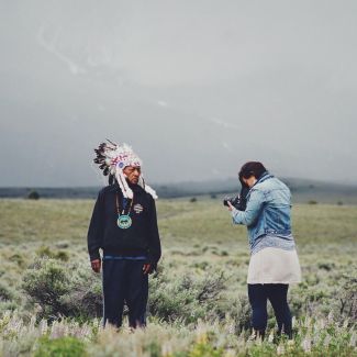 Photographer Matika Wilbur taking a picture of a Native American man wearing a traditional head covering.