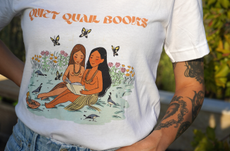 Photo of a slim, tan person's torso wearing a Quiet Quail Books t-shirt and with intricate tattoos on their left arm