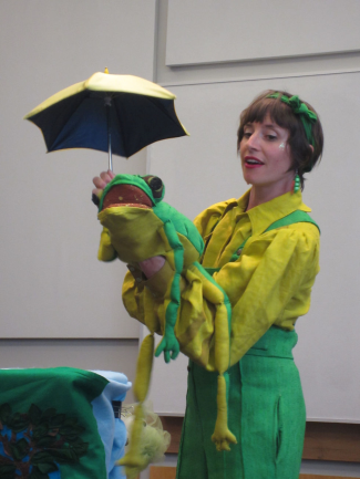 photo of Risa from Jelly Jam time wearing all green holding a large frog puppet and holding a small umbrella over the frog's head