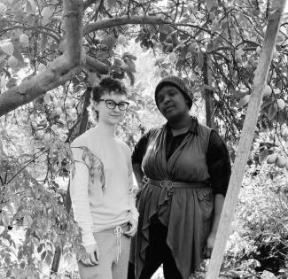 Sepia toned photo of Alyssa, a white person with glasses wearing white and gray, and Sej, a Black person wearing a beanie, a collared shirt, and a belted drapey top layer.