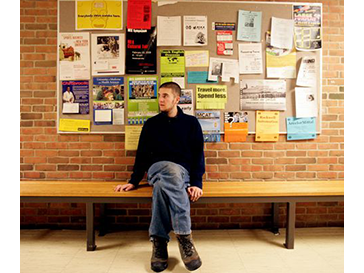A young white man sitting on a bench in front of a wall covered in flyers.