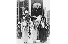 Three women in traditional clothes holding a banner advocating for suffrage.