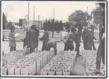 Scanned photograph in a newspaper, showing rows of grocery bags in a public park and about a dozen people milling around to pick them up