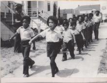 Black and white photograph of a group of school children marching down a residential street in unison. The children are wearing a uniform of black pants, white shirts, and black berets.