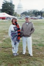 An older man and woman in a field smiling at a camera. In the distance, the United States capital is visible