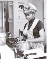 Black and white photograph of a young woman in an apron cooking in a kitchen