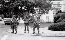 Black and white photograph of three national guardsmen in full gear reading a newspaper on a residential street corner