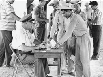 Man at a table outside with another man behind it with men in background