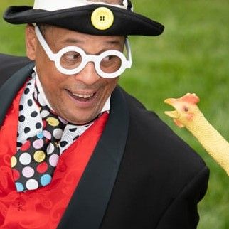 Unique Derique wearing large white glasses, a black jacket and a red vest, smiles at a rubber chicken. 